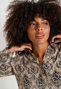 Blusa color tostado con estampado floral negro, con cuello abotonado y detalles fruncidos en las mangas, llevada por una persona con cabello rizado.