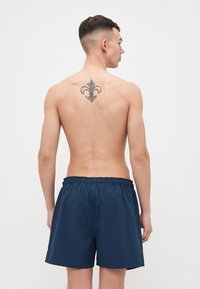 Mørkeblå svømmedrakter med elastisk linning, med en glatt tekstur og sidelommer, båret av en mannlig modell sett bakfra.