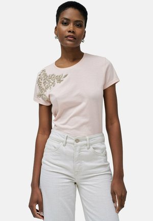 Camiseta de algodón de color rosa claro con intrincados bordados florales de cuentas en el hombro izquierdo. Combinada con jeans de tiro alto de color blanco roto.
