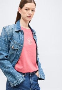 Γυναίκα που φοράει μπλε τζιν μπουφάν με εκτυπωμένο κείμενο πάνω σε ροζ πουκάμισο που γράφει "Πάντα κυνηγώντας τα ηλιοβασιλέματα," σε συνδυασμό με μπλε τζιν παντελόνι.