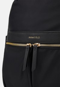 Sac à dos en nylon noir avec une texture lisse, des fermetures éclair dorées et un accent en cuir noir portant l'inscription "ANNA FIELD" en relief.