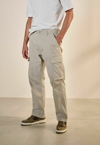 Pantalones cargo beige de algodón con múltiples bolsillos y un corte relajado. Combinados con zapatillas en marrón oscuro y crema sobre un suelo liso.