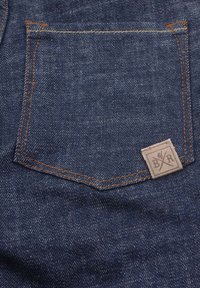 Tumma sininen farkkukangas sisältää läpällisen taskun, jossa on ruskea ommel ja brändilogo reunassa. Kankaan pinta on sileä ja kudos on näkyvissä.