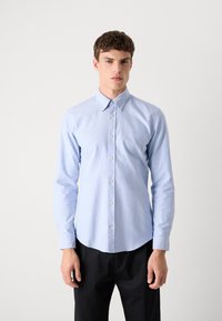 Šviesiai mėlyna ilgomis rankovėmis marškiniai su mygtukų apykakle, subtilia tekstūra ir maža išsiuvinėta logo kairiajame krūtinės pusėje. Ilgos rankovės turi rankogalius.