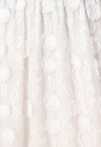 Witte stof met ingewikkelde bloemenborduursels, voorzien van textuurrijke pompons en zwierende bladerenpatronen. Zacht en delicaat uiterlijk.