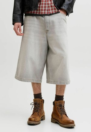 Person iført lysegrå knælange cowboyshorts, brune snørebåndsstøvler med mørke sokker, rød ternet skjorte og sort læderjakke.