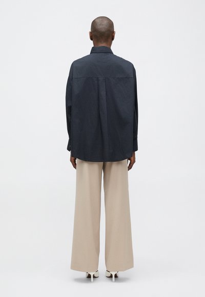 Osoba v voľnej tmavomodrej košeli, širokých béžových nohaviciach a bielych podpätkových topánkach, stojí chrbtom k jednoduchému pozadiu.