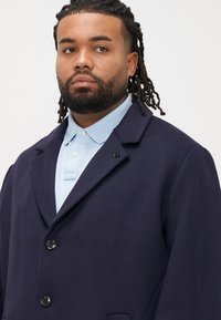 Cappotto blu navy in misto lana con collo sciallato, tre bottoni neri e tasche laterali, indossato sopra una polo azzurro chiaro.