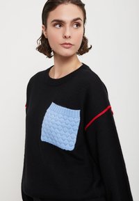 Maglione nero in maglia con una tasca di un azzurro chiaro testurizzato. Presenta dettagli rossi sulle spalle. Collo rotondo e vestibilità ampia.