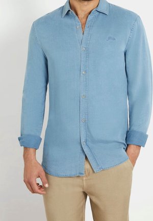 Man wearing light blue button-up denim shirt with rolled sleeves and beige pants, one hand in pocket, standing against plain background.