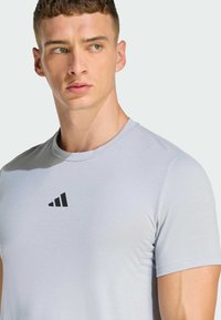 Camiseta atlética gris claro con mangas cortas y cuello redondo. Presenta un pequeño logo negro en el pecho. Tela suave y texturizada.