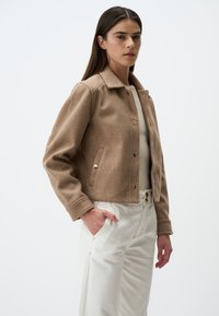Veste cropped en mélange de laine marron avec un col, des boutons dorés et des poches latérales, associée à un pantalon taille haute de couleur crème.