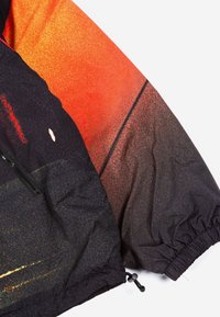 Vēja pārklājs ar gradienta dizainu oranžā un melnā krāsā. Materiāls ir viegls, ar elastīgām aprocēm un ar rāvējslēdzēju aprīkotu kabatu.