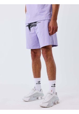 Hombre vistiendo pantalones cortos lavanda con diseño gráfico negro, calcetines blancos con logo y zapatillas blancas Nike, de pie sobre un fondo blanco.