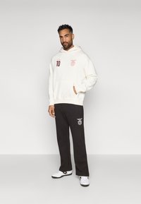 Sudadera blanca con capucha y bolsillo delantero, con un logo burdeos, combinada con pantalones de chándal negros que tienen un logo blanco. El modelo lleva zapatillas blancas.
