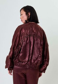 Bordo satino bomberis su susiraukšlėjimu nugaros dalyje ir pūstomis rankovėmis, su elastingu juosmeniu ir rankogaliais.
