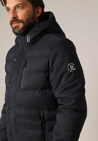 Homme avec une barbe portant une veste rembourrée noire avec capuche, avec des poches zippées sur la poitrine et les côtés, se tenant devant un fond uni.