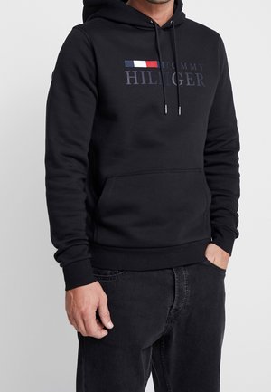 Mężczyzna noszący czarną bluzę z kapturem Tommy Hilfiger z logo i wzorem flagi na piersi oraz czarne jeansy, z jedną ręką w kieszeni.