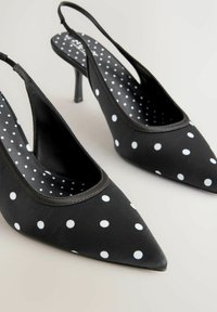 Pantofi cu toc slingback negri cu buline, cu vârf ascuțit, având un finisaj lucios, toc subțire și modele circulare albe pe suprafață.