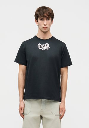 Νεαρός με κοντά καστανά μαλλιά, που φοράει μαύρο μπλουζάκι με λευκό στυλιζαρισμένο κείμενο "GCDSWEAR" και ανοιχτό μπεζ παντελόνι, στέκεται μπροστά σε απλό φόντο.