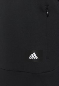 Felpa nera con tessuto testurizzato, tasca con zip e un'etichetta nera con il logo bianco di Adidas. Design e forma semplici.