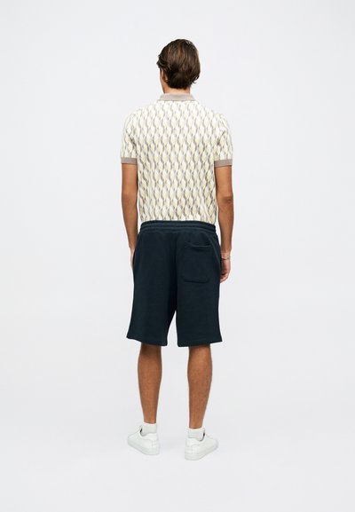 Мъж, облечен с поло с шарени къси ръкави, тъмни къси панталони до коляното с джоб отзад, бели маратонки, стои с лице към отсрещната страна на обикновен фон.