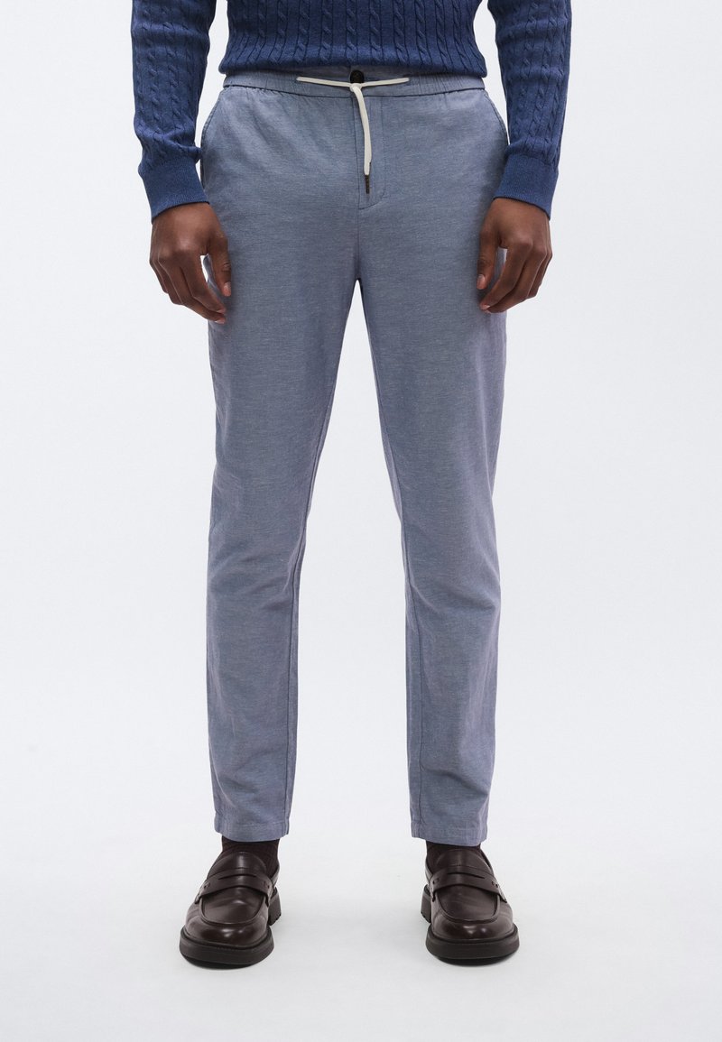 Ανοιχτό μπλε, στενά παντελόνια με ελαστική μέση με κορδόνι, φτιαγμένα από υφασμάτινο υλικό με υφή, συνδυασμένα με σκούρα καφέ παπούτσια χωρίς κορδόνια.