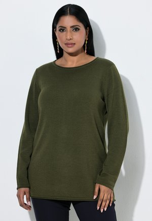 Mujer con el pelo largo y negro, que lleva una camisa verde de manga larga y pendientes colgantes de colores, de pie contra una pared blanca.