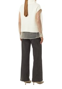 Vrouw met lang haar draagt een mouwloze witte gebreide vest over een doorschijnende blouse met lange mouwen, losse donkergrijze broek en beige kitten heels.