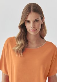 Oranžový top s krátkym rukávom a širokým výstrihom, s béžovým lemovaním. Mäkka tkanina s hladkou textúrou, voľne padá cez ramená.