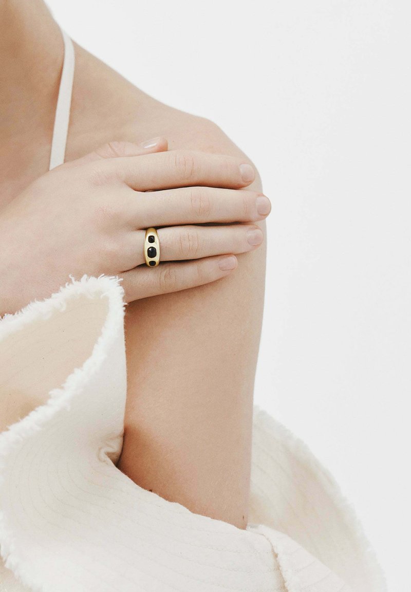 Anillo de oro con dos piedras negras, con un diseño liso y redondeado. Mano apoyada en el hombro, contra una tela suave y texturizada.
