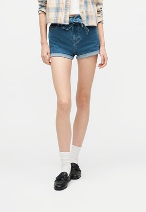 Kobieta w krótkich spodenkach dżinsowych z podwiniętymi brzegami i zawiązanym w talii paskiem, białych skarpetkach, czarnych mokasynach oraz w kraciastej koszuli na białym topie.