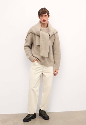 Maglione beige a maglia sovrapposto con una sciarpa abbinata, abbinato a pantaloni crema chiara e stivali stringati verde scuro su un pavimento grigio.