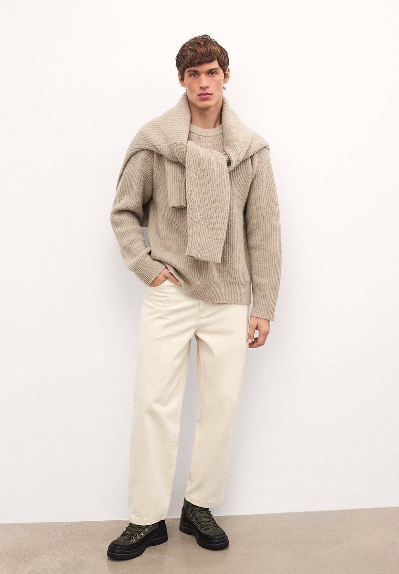 Maglione beige a maglia sovrapposto con una sciarpa abbinata, abbinato a pantaloni crema chiara e stivali stringati verde scuro su un pavimento grigio.