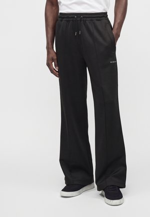 Persona che indossa pantaloni neri larghi con coulisse e sneakers nere con suole bianche, in piedi davanti a uno sfondo semplice.