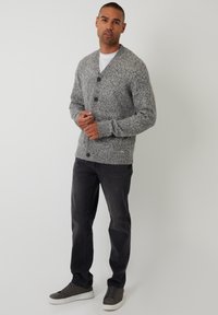 Cardigan gris de punto con cuello en V, dos botones y puños acanalados. Lleva puesto sobre una camisa blanca, combinado con jeans oscuros y zapatillas grises.