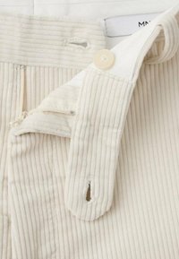 Pantalones blancos de pana con textura acanalada vertical, cierre frontal con botón y cremallera. Incluyen un lazo para colgar y una etiqueta con el nombre de la marca.