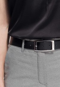 Cinturón de cuero negro con hebilla rectangular plateada, usado con pantalones de espiga gris y una blusa de satén negra.