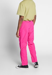 Man med ljusrosa byxor, beige t-shirt och svarta Converse-sneakers, stående mot en enkel vit bakgrund.