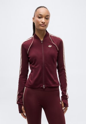 Kobieta ubrana w bordową kurtkę sportową Adidas i dopasowane legginsy z kremowymi paskami, stojąca na tle jednolitego jasnego tła.