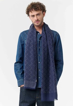 Man with curly hair wearing dark denim shirt and jeans, draped navy patterned scarf, standing against plain light background.