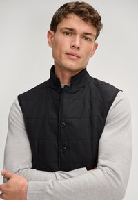 Czarna pikowana kamizelka z wysokim kołnierzem, zapinana na dwa guziki, noszona na jasnoszarej koszuli z długim rękawem. Gładka faktura materiału.