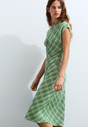Vrouw met lang haar die een mouwloze groene geruite midi-jurk draagt, kijkt opzij tegen een effen lichte achtergrond.