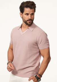 Helles rosa, gestricktes Polohemd mit kurzen Ärmeln und V-Ausschnitt, strukturiertem Stoff, gerippten Bündchen und Saum. Getragen mit weißen Hosen und Accessoires.