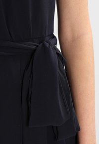 Marinblå klänning i texturerad tyg, med ett knutet bälte i midjan och flödande tyg som skapar en mjuk silhuett.