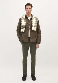 Zelená bavlněná košile-jacket, bílý pruhovaný pletený svetr přehozený přes ramena, grafické tričko, olivové kalhoty a černé boty, které nosí modelka.