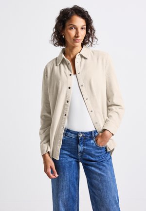 Vrouw in een beige corduroy overhemd over een witte top en blauwe jeans, staande met één hand in de zak tegen een witte achtergrond.