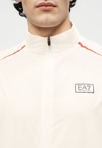 Άνδρας που φοράει λευκό μπουφάν με φερμουάρ, με κόκκινες ρίγες στους ώμους και το λογότυπο EA7 Emporio Armani στο στήθος.