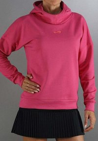 Mujer vestida con una sudadera rosa brillante con un pequeño logo naranja y una minifalda negra plisada, de pie con una mano en la cadera.