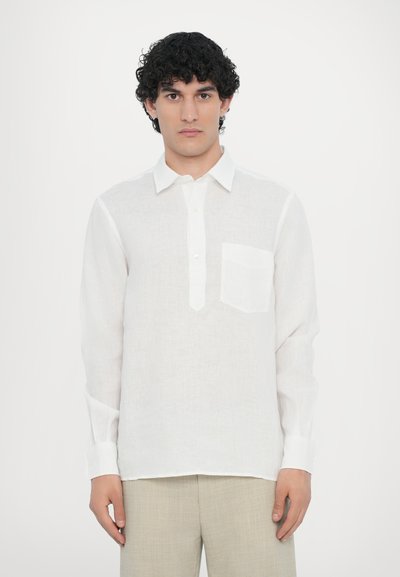 Chemise blanche en lin à manches longues avec un col boutonné et une poche sur le devant, présentant une coupe décontractée et des manches retroussées. Texture lisse et légère.